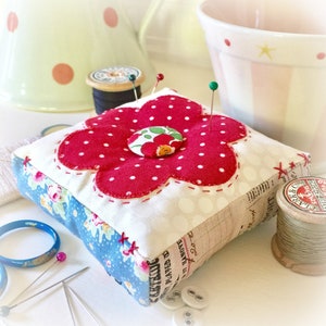 May include: A red and white polka dot flower-shaped pincushion with a smaller red button in the center. The pincushion is made of fabric and has a blue and white patterned fabric on the sides. There are several sewing pins stuck in the pincushion.