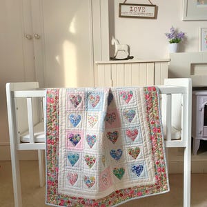 Puede incluir: Una cuna blanca con una colcha hecha a mano. La colcha presenta parches en forma de corazón con diseños florales en varios colores, incluyendo rosa, azul y verde. El borde de la colcha tiene un diseño floral. Un letrero encima de la cuna dice "LOVE MAKES IT ALL WORTH WHILE".