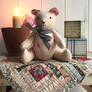 May include: A handmade patchwork teddy bear with a black and white gingham ribbon around its neck. The bear is sitting on a quilt with a red, white, and black pattern. The bear is surrounded by a candle, a flower, and a wooden block with the alphabet printed on it.