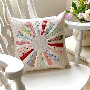 May include: A decorative square pillow with a colorful, circular patchwork design. The patchwork features various patterned fabrics in shades of red, pink, blue, and green. The pillow is on a white chair.