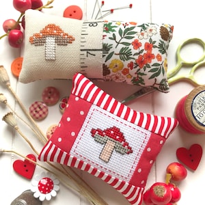 Puede incluir: Dos pequeños alfileteros a rayas rojas y blancas con setas bordadas. Un alfiletero tiene una tela floral en la parte posterior.