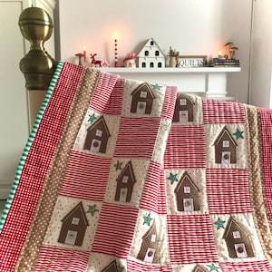 May include: A red and white striped quilt with gingerbread house appliques. The quilt is folded on a bed with a green and white gingham border.