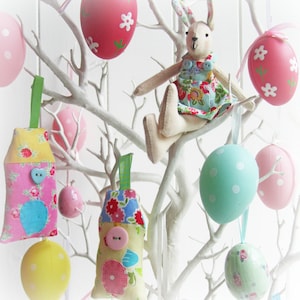 May include: A white branch tree decorated with pastel colored Easter eggs, a bunny rabbit doll, and fabric ornaments. The ornaments are shaped like houses and eggs and have floral patterns.