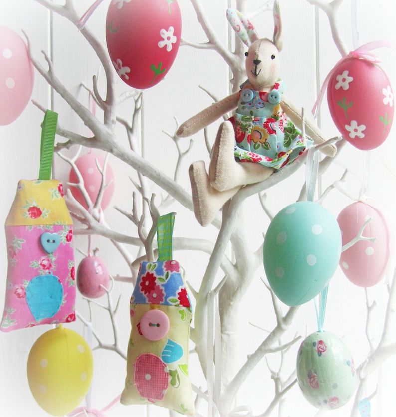 May include: A white branch tree decorated with pastel colored Easter eggs, a bunny rabbit doll, and fabric ornaments. The ornaments are shaped like houses and eggs and have floral patterns.