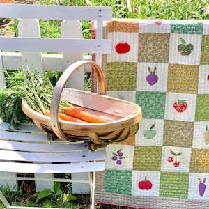May include: A white metal chair supports a wooden basket brimming with carrots. A patchwork quilt with fruit and vegetable motifs is draped nearby. The quilt displays squares with images of apples, cherries, and beetroot. The scene is set outdoors in a garden.