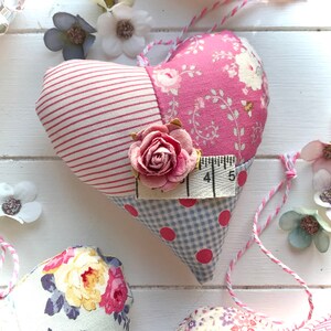 May include: A pink and blue patchwork heart-shaped pillow with a floral print and a small fabric rose. The pillow has a white and pink striped fabric on one side and a blue and white gingham fabric on the other side. The pillow is decorated with a small fabric rose and a measuring tape.