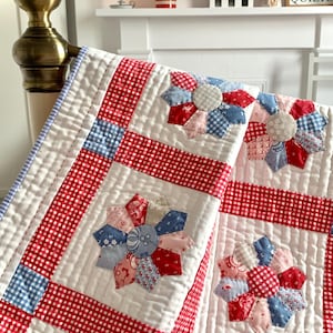 Puede incluir: Un edredón hecho a mano con fondo blanco, bordes de cuadros rojos y diseños florales de patchwork en rojo, azul y rosa. El edredón está colocado sobre un marco de cama, mostrando sus intrincadas costuras y colores vibrantes.