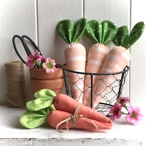 Può includere: Tre carote di stoffa con foglie verdi a pois bianchi in un cesto di filo metallico. Due carote di stoffa aggiuntive sono legate insieme con uno spago e sono appoggiate su una superficie bianca. Sullo sfondo ci sono dei fiori rosa.