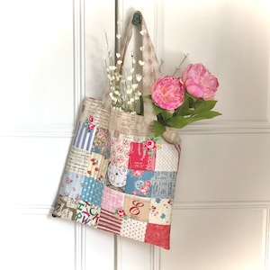 Può includere: Una borsa a tracolla in patchwork con stampa floreale e alfabetica è appesa a una porta bianca. La borsa è piena di fiori bianchi e peonie rosa.