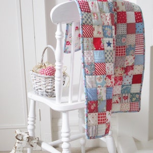 May include: A white wooden chair with a patchwork quilt draped over the back. The quilt is made of squares of red, blue, and white fabric with various patterns. The chair is sitting in a room with white walls and a white rocking horse in the foreground.