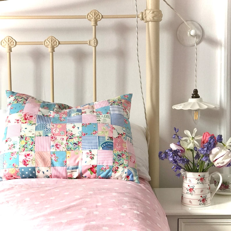 May include: A white metal bed frame with a pink and blue patchwork pillow on top. The bed has a pink polka dot duvet cover. A floral patterned ceramic pitcher with a bouquet of purple and white flowers sits on a white nightstand next to the bed.