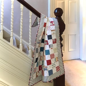 May include: A red, white and blue patchwork quilt with a striped border. The quilt is draped over a wooden banister.
