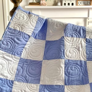 May include: A light blue and white patchwork quilt with a spiral quilted design. The quilt is displayed over a bed frame. The squares are arranged in a checkerboard pattern, showcasing the handmade quality and texture of the quilt.