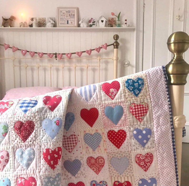 May include: Two white quilts with a patchwork design of red, blue, and white hearts. The quilts are on a white bed with a pink pillow and a white headboard. A pink heart garland is hanging above the headboard.