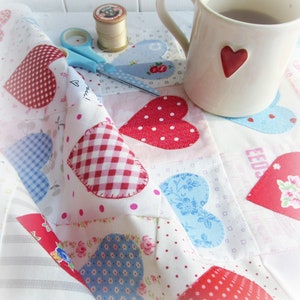 May include: A white fabric quilt with a patchwork design featuring red and blue hearts. The quilt is made of various fabrics with different patterns, including gingham, polka dots, and floral prints. A mug of tea and a spool of thread are visible in the background.