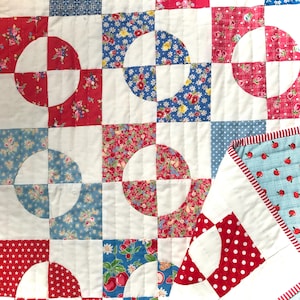 Op de afbeelding: Een wit quilt met een patchwork-ontwerp met rode, blauwe en witte bloemenstof. Het quilt is gemaakt van vierkanten met cirkels die uit het midden zijn geknipt. Het quilt is gestikt met wit garen.