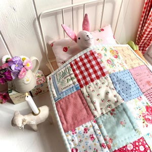 May include: A white doll bed with a pink and white floral patterned pillow and a patchwork quilt. The quilt is made of various fabrics with floral, checkered, and striped patterns in red, blue, and white. The quilt has the text "Lemons are rich!" printed on it.