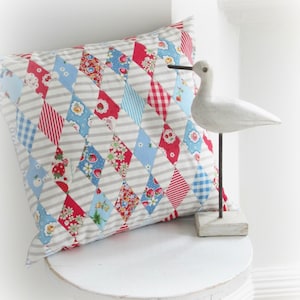May include: A white pillow with a diamond pattern in red, blue, and white fabric. The pillow is sitting on a white stool with a white wooden bird figurine on the right side of the pillow.