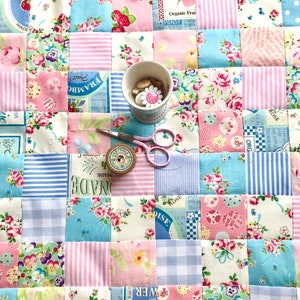 May include: A patchwork quilt with a variety of floral patterns in pink, blue, and white. The quilt is made up of squares, and each square has a different pattern. There are also some buttons and a spool of thread on the quilt.