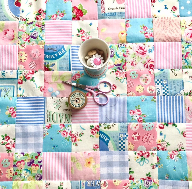 May include: A patchwork quilt with a variety of floral patterns in pink, blue, and white. The quilt is made up of squares, and each square has a different pattern. There are also some buttons and a spool of thread on the quilt.