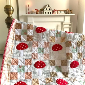 Op de afbeelding: Een wit en groen patchwork-deken met rode en witte paddenstoel-applicaties. Het deken ligt op een bed met een rood-witte ruitjesrand.