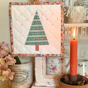 May include: A square quilted potholder featuring a white center with a green floral patterned Christmas tree applique, surrounded by a colorful floral border and a matching hanging loop, accented with small orange embroidered star details.