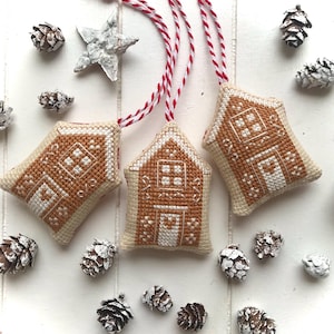 May include: Three gingerbread house ornaments with white string hangers. The houses are made of beige fabric with brown gingerbread details and white icing accents. The houses are hanging against a white wood background.