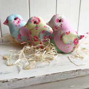 Op de afbeelding: Drie handgemaakte stoffen vogels met bloemenpatronen. De vogels bevinden zich in een nest van versnipperd papier. De vogels zijn lichtblauw, geel en roze.
