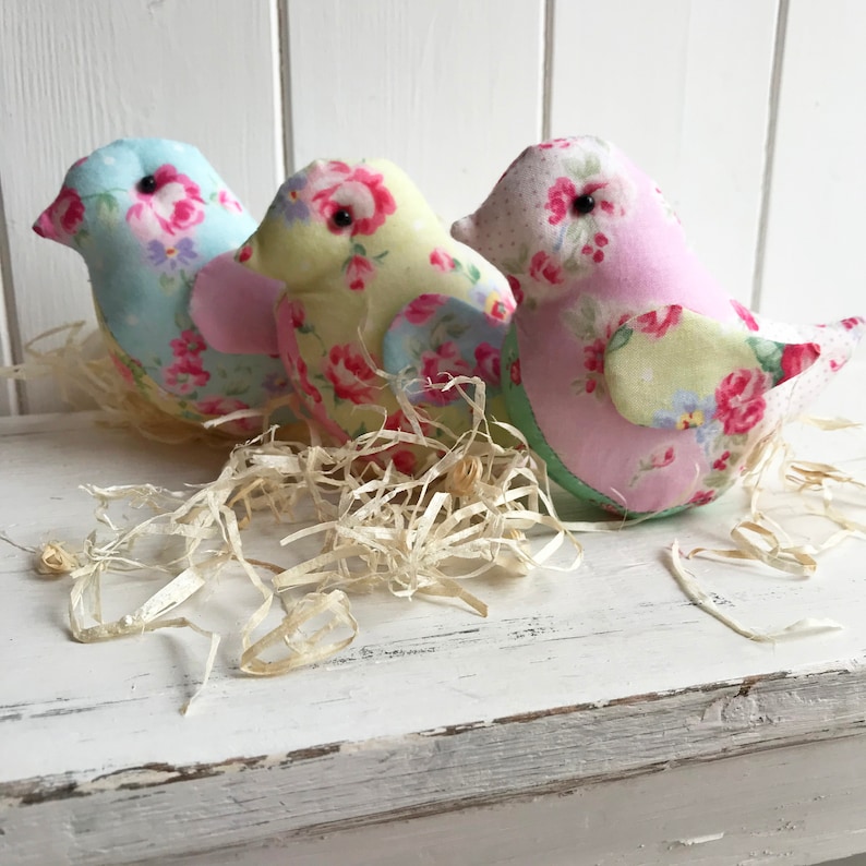 Op de afbeelding: Drie handgemaakte stoffen vogels met bloemenpatronen. De vogels bevinden zich in een nest van versnipperd papier. De vogels zijn lichtblauw, geel en roze.