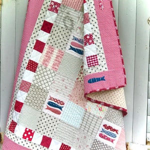 Puede incluir: Una colcha de retazos con un esquema de color rojo, blanco y azul. La colcha está hecha de varias telas con diferentes patrones, incluyendo estrellas, rayas y peces. La colcha está colgada en una pared blanca.