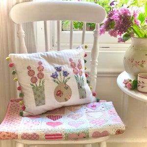 Puede incluir: Una silla blanca con un cojín blanco con un diseño floral. El cojín tiene tres jarrones con flores de diferentes colores. La silla está sentada frente a una ventana con vista a un jardín.