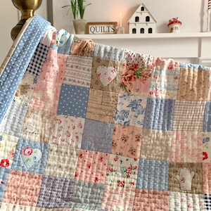 以下が含まれることがあります： パッチワークのキルト。パステルピンク、ブルー、クリーム色の花柄、水玉模様、柄物の生地を使用。ハート型とミトン型のアップリケ付き。縁は水色のギンガムチェック柄。