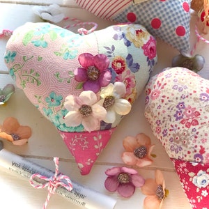 May include: A pink and blue patchwork heart-shaped pillow with floral fabric and pink trim. The pillow is decorated with white and purple fabric flowers.