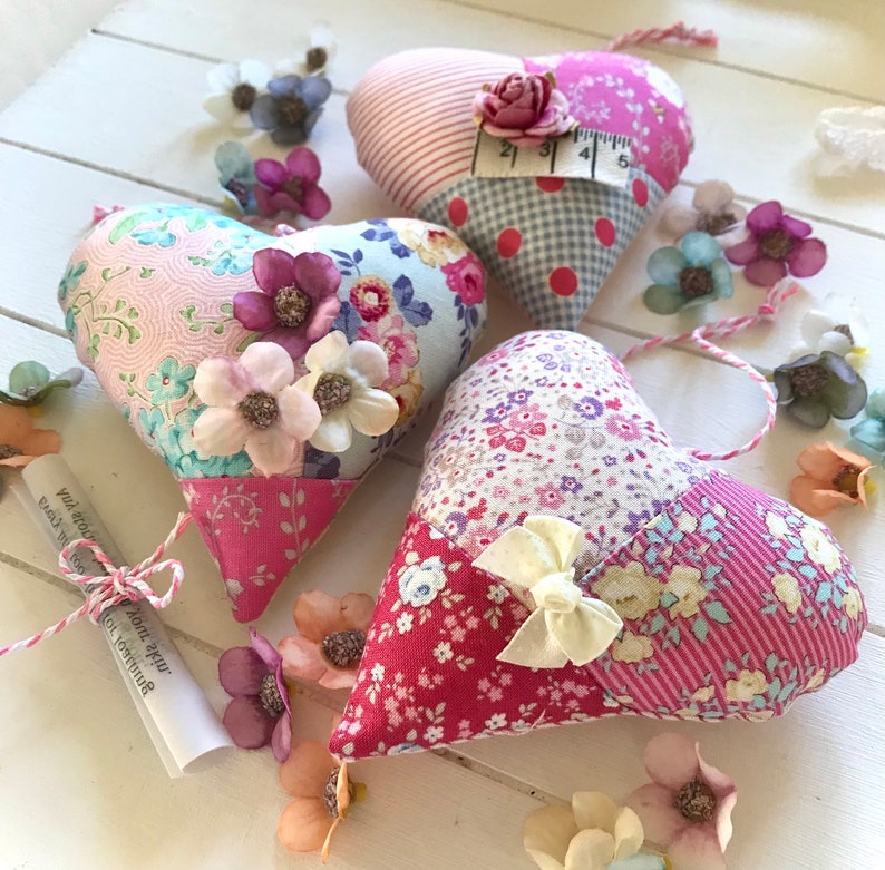 May include: Three pink and white patchwork fabric heart ornaments with floral embellishments and a white ribbon bow. The hearts are hanging from white string.