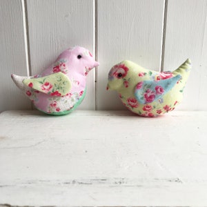 Op de afbeelding: Twee handgemaakte stoffen vogels met bloemenpatronen. De vogel links is roze met groene accenten en de vogel rechts is geel met blauwe accenten.