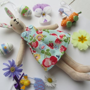 以下が含まれることがあります： 白い背景に、赤いチェリーがついた青と白の花柄のドレスを着た、手作りの布製のウサギのぬいぐるみ。長い垂れ下がった耳があり、様々なイースターの飾り付けが施されています。