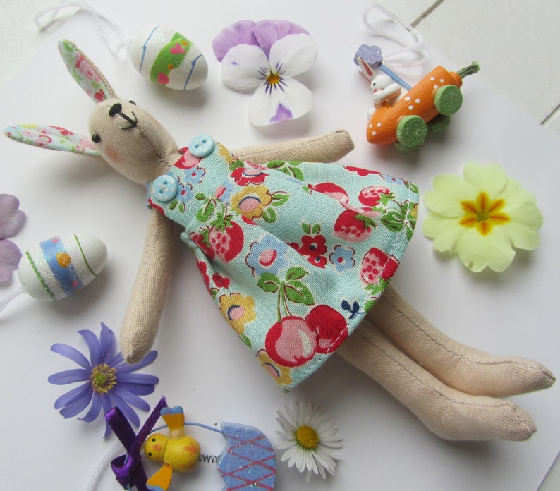 May include: A handmade fabric bunny doll wearing a blue and white floral dress with red cherries. The doll has long, floppy ears and is lying on a white surface with various Easter decorations.