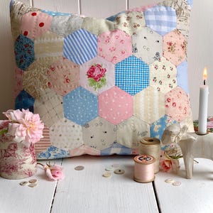 Puede incluir: Un cojín de patchwork con una variedad de telas estampadas en tonos rosa, azul y crema. El cojín presenta un motivo central de rosa y está rodeado de carretes de hilo, botones y un pequeño jarrón con flores.