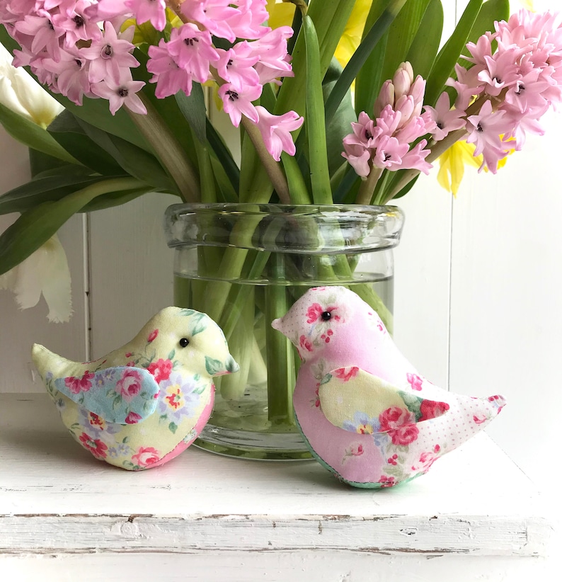 Op de afbeelding: Twee handgemaakte vogels van stof, een gele met blauwe accenten en een roze met gele accenten, die op een wit oppervlak zitten voor een glazen vaas gevuld met roze en gele hyacinten.