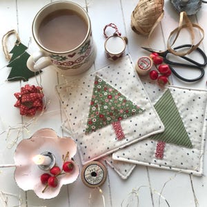 以下が含まれることがあります： 緑と赤のクリスマスツリーのデザインが施された白い布製のコースターが2つ。コースターは、白い木製の表面に置かれ、コーヒーカップ、糸巻き、その他のクリスマスの装飾品が置かれている。