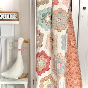 May include: A white quilt with a floral pattern. The quilt is made up of hexagonal shapes, each with a different floral design. The quilt is hanging on a white door with a white goose figurine in the foreground. The sign above the goose reads "QUILTS".