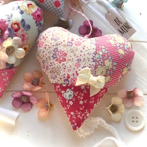 May include: A patchwork heart-shaped pillow with a floral print and a white bow. The pillow is made of different fabrics and has a pink, blue, and yellow color scheme. The pillow is sitting on a white surface with a tag that says "Be Mine".
