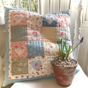 Puede incluir: Una almohada de patchwork con un borde de lunares azules y blancos. La almohada está hecha de varias telas con estampados florales y corazones. La almohada está sentada sobre una superficie blanca con una pequeña planta en maceta delante.