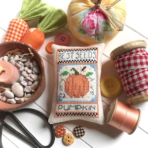 May include: A white pillow with a cross-stitch design of a pumpkin seed packet. The packet is labeled "Best Seeds Pumpkin". The pillow is surrounded by fall-themed decorations, including a fabric pumpkin, a spool of red and white gingham ribbon, and a spool of orange thread.