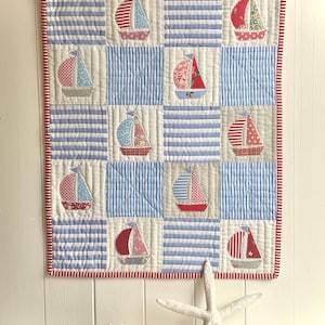 Könnte beinhalten: Ein Wandbehang mit maritimem Thema, der ein Patchwork-Design aufweist. Er zeigt blau-weiß gestreifte Quadrate und applizierte Segelboote mit roten, rosa und blauen Segeln mit verschiedenen Mustern. Der Quilt ist mit einem rot-weiß gestreiften Stoff eingefasst.