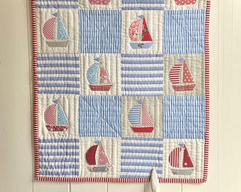 May include: A nautical-themed wall hanging featuring a patchwork design with blue and white striped squares and appliqued sailboats. The sailboats have red, pink, and blue sails with various patterns. The quilt is bordered with a red and white striped fabric.