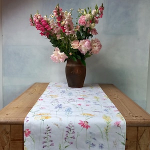 Könnte beinhalten: Ein Holztisch mit einem floralen Tischläufer und einer Blumenvase. Der Tischläufer ist weiß mit einem bunten Wildblumenmuster. Die braune Vase enthält rosa und rote Blumen.