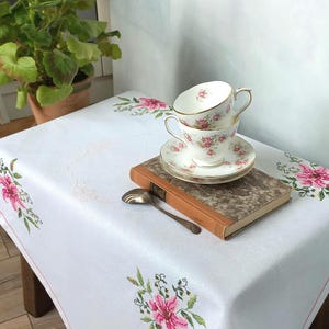 Op de afbeelding: Een witte tafelkleed met roze bloemenborduurwerk, twee gestapelde kopjes en schotels, een vintage boek en een lepel. Een potplant met roze bloemen staat op de achtergrond. De kopjes en schotels hebben een roze bloemenpatroon.