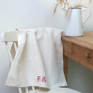 May include: Cream-colored linen towel draped over a white chair. The towel features red embroidered initials. A white enamel pitcher with a black rim and wheat stalks sits on a wooden table, creating a rustic, vintage aesthetic.