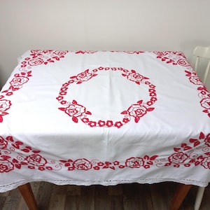 Pode incluir: Toalha de mesa quadrada branca com bordado de rosas vermelhas. A toalha de mesa apresenta uma borda de rosas e uma grinalda central de rosas. As bordas são finalizadas com uma delicada borda de renda. Uma peça decorativa para uma mesa.